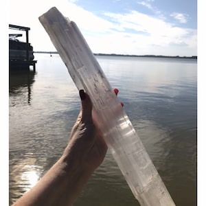 Pode incluir: Um grande cristal de selenita transparente, também conhecido como varinha de selenita, é segurado na mão contra um fundo de lago e céu. O cristal é liso e tem uma textura estriada.