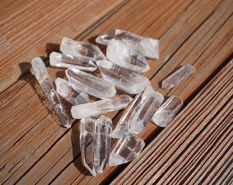 May include: A collection of clear quartz crystal points on a wooden surface. The crystals are various sizes and shapes, with some having a pointed tip and others having a more rounded tip.