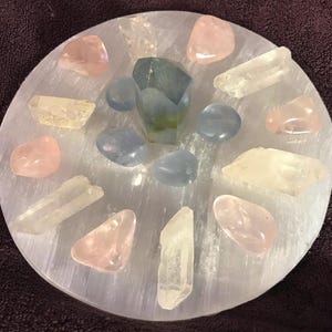 May include: A selenite charging plate with a variety of crystals, including clear quartz, rose quartz, and blue calcite. The crystals are arranged in a circle around a larger, green and blue crystal in the center.