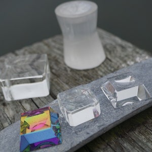 May include: Four clear glass display stands of varying sizes and shapes, with a rainbow prism on a gray slate surface.