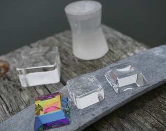 Puede incluir: Cuatro soportes de exhibición de vidrio transparente de diferentes tamaños y formas, con un prisma arcoíris sobre una superficie de pizarra gris.