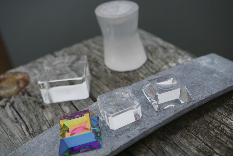 May include: Four clear glass display stands of varying sizes and shapes, with a rainbow prism on a gray slate surface.