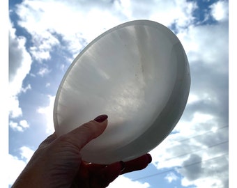 Puede incluir: Un plato de carga de selenita blanco, un plato de cristal redondo y liso, sostenido contra un cielo azul con nubes blancas.