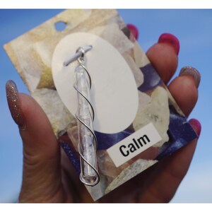 Pu&ograve; includere: Un ciondolo avvolto in filo d'argento con cristalli trasparenti all'interno di un tubo di vetro. Il ciondolo &egrave; su una carta bianca con la parola "Calm" stampata su di essa.