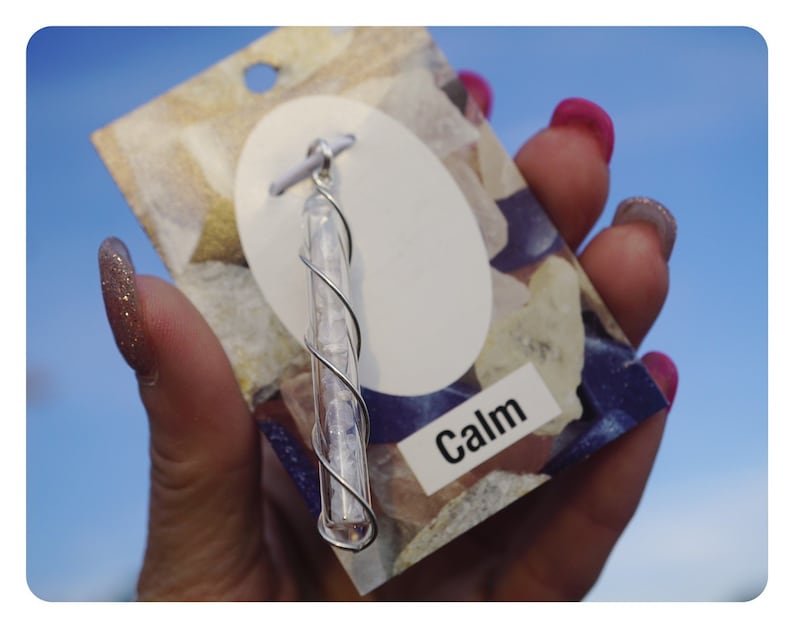 Pu&ograve; includere: Un ciondolo avvolto in filo d'argento con cristalli trasparenti all'interno di un tubo di vetro. Il ciondolo &egrave; su una carta bianca con la parola "Calm" stampata su di essa.