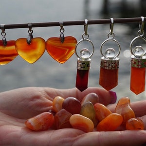 May include: A collection of gemstone pendants and tumbled stones. The pendants include heart-shaped and rectangular designs in shades of orange and red, suspended from silver-colored metal. A hand holds a variety of polished, orange-toned tumbled stones.
