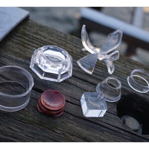 May include: A set of clear acrylic display stands for showcasing small objects. The set includes a variety of shapes and sizes, including a round ring, a square base, a cone, and a star-shaped stand. There is also a small wooden stand.