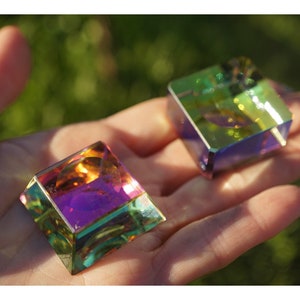 May include: Two iridescent glass cubes, one orange, purple, and green, the other green, blue, and yellow, are held in a hand against a green background.