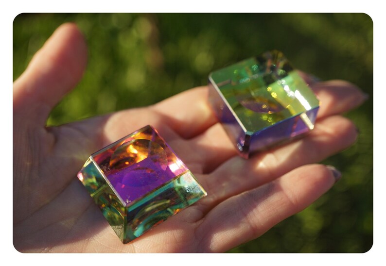 May include: Two iridescent glass cubes, one orange, purple, and green, the other green, blue, and yellow, are held in a hand against a green background.