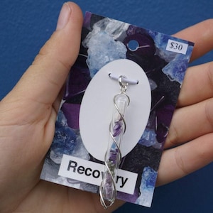 May include: A clear glass pendant necklace with a silver wire wrap design, filled with purple and clear crystals. The pendant hangs from a silver chain. The necklace is displayed on a card with the word "Recovery".