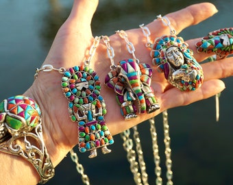 May include: A hand displays a collection of silver necklaces and a ring, each adorned with colorful, mosaic-style gemstones. The pendants feature Native American figures, an elephant, and a ring with a skull design. The jewelry showcases a vibrant mix of turquoise, red, and green stones.