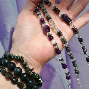 K&ouml;nnte beinhalten: Eine Sammlung von Edelsteinschmuck, darunter Armb&auml;nder und Halsketten. Die Armb&auml;nder haben dunkelgr&uuml;ne und schwarze Perlen, w&auml;hrend die Halsketten eine Vielzahl von lila, klaren und gr&uuml;nen Steinen zeigen. Der Schmuck wird vor einem weichen, lila Hintergrund pr&auml;sentiert.