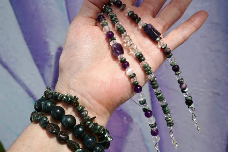 K&ouml;nnte beinhalten: Eine Sammlung von Edelsteinschmuck, darunter Armb&auml;nder und Halsketten. Die Armb&auml;nder haben dunkelgr&uuml;ne und schwarze Perlen, w&auml;hrend die Halsketten eine Vielzahl von lila, klaren und gr&uuml;nen Steinen zeigen. Der Schmuck wird vor einem weichen, lila Hintergrund pr&auml;sentiert.
