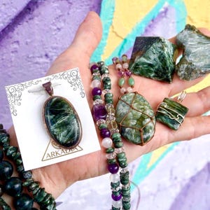 K&ouml;nnte beinhalten: Eine Hand pr&auml;sentiert verschiedene gr&uuml;ne und violette Edelsteinschmuckst&uuml;cke und Rohsteine. Zu den Artikeln geh&ouml;ren ein Anh&auml;nger auf einer Karte mit dem Text "ARKADIA", Perlenketten, Ohrringe und rohe Mineralproben. Die Edelsteine haben eine Mischung aus gr&uuml;nen und violetten Farbt&ouml;nen.