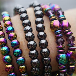 Puede incluir: Cinco pulseras hechas de cuentas negras iridiscentes. Las pulseras est&aacute;n apiladas en la mu&ntilde;eca de una persona. Las cuentas son redondas y tienen un brillo met&aacute;lico.