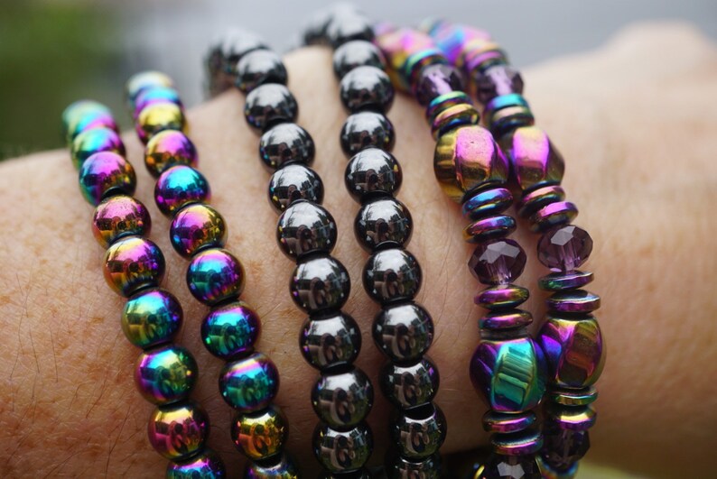 Puede incluir: Cinco pulseras hechas de cuentas negras iridiscentes. Las pulseras est&aacute;n apiladas en la mu&ntilde;eca de una persona. Las cuentas son redondas y tienen un brillo met&aacute;lico.