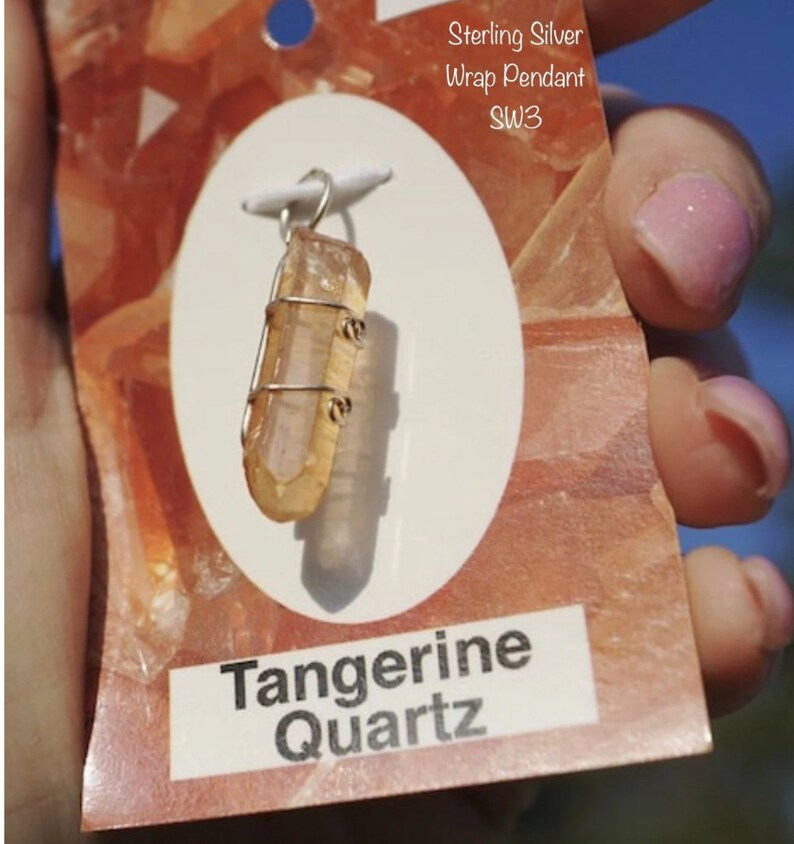 May include: A sterling silver wrap pendant featuring a tangerine quartz crystal. The pendant is wrapped in silver wire and has a loop for attaching a chain. The tag reads "Sterling Silver Wrap Pendant SW3 Tangerine Quartz".