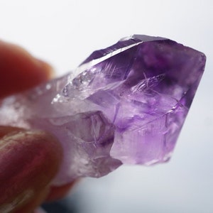 May include: A purple amethyst crystal with a pointed tip and clear facets. The crystal is translucent and has a light purple hue.