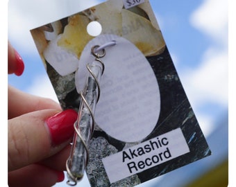 May include: A silver wire wrapped around a clear glass tube filled with small gray stones. The tube is hanging from a white string. The tag attached to the tube reads "Akashic Record" and is priced at $30.