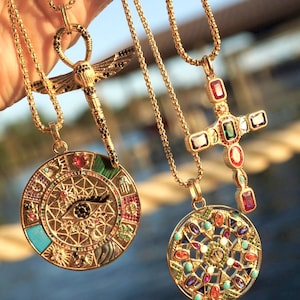 May include: Four gold necklaces with different pendants. The pendants are a dragonfly, a circle with an eye design, a circle with a geometric design, and a cross. All pendants are decorated with colorful gemstones.