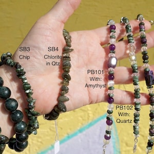 K&ouml;nnte beinhalten: Eine Auswahl an Edelsteinarmb&auml;ndern mit runden, Chip- und Nugget-Designs, mit Amethyst-, Quarz- und Charoit-Akzenten. Die Armb&auml;nder sind mit Texten wie "SB2 12MM Round" und "PB101 With: Amythyst" beschriftet.