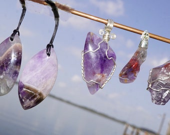 May include: Five amethyst crystal pendants, some wrapped in silver wire, hanging from a copper rod. The pendants are various shapes and sizes, with some having a purple and white color.