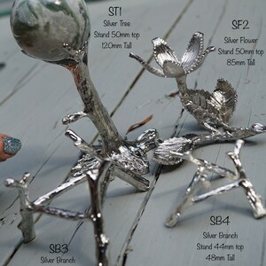 May include: Four silver metal decorative stands for displaying small objects. The stands are shaped like a tree, a flower, and two branches. The stands are on a light wood surface.  STI Silver Tree Stand 50mm top 120mm Tall. SF2 Silver Flower Stand 50mm top 85mm Tall. SB3 Silver Branch Stand 60mm top 66mm Tall. SB4 Silver Branch Stand 44mm top 48mm Tall.
