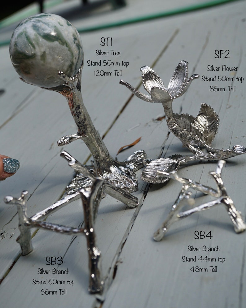 May include: Four silver metal decorative stands for displaying small objects. The stands are shaped like a tree, a flower, and two branches. The stands are on a light wood surface.  STI Silver Tree Stand 50mm top 120mm Tall. SF2 Silver Flower Stand 50mm top 85mm Tall. SB3 Silver Branch Stand 60mm top 66mm Tall. SB4 Silver Branch Stand 44mm top 48mm Tall.