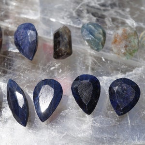 Könnte beinhalten: Eine Sammlung von birnenförmigen blauen Saphiren, einige mit silberner Rückseite, und andere Edelsteine in verschiedenen Blautönen und Grüntönen.