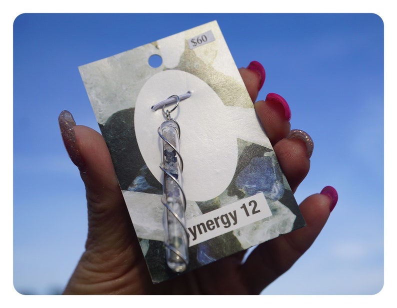 Puede incluir: Un colgante de plata con un tubo de vidrio transparente lleno de peque&ntilde;as piedras azules y blancas. El colgante est&aacute; en una tarjeta blanca con el texto "Synergy 12" y una etiqueta de precio de 60 &euro;.
