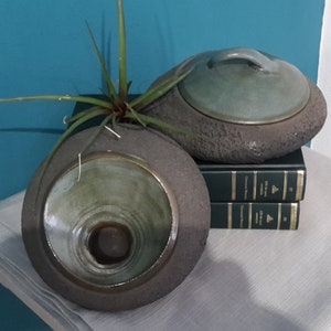 May include: Two ceramic vessels with a stone-like texture, one open and the other with a lid. The open vessel holds a small plant. Both have a green interior and sit on a stack of books and a linen cloth.