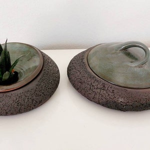 May include: Two ceramic planters with a unique design. One contains a succulent plant. The planters have a dark brown, textured base and a teal-colored interior. One planter has a lid with a handle. The planters are suitable for home decor.