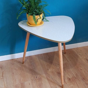 Può includere: Un tavolino triangolare bianco con un piano in laminato bianco e un bordo dorato. Il tavolino ha tre gambe in legno con punte dorate. Una pianta di felce in vaso è appoggiata sul tavolino.