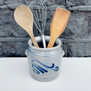 以下が含まれることがあります： 渦巻き模様の青と白のセラミック製の食器入れ。入れ物には木製のさじ、木製のヘラ、ワイヤー製の泡立て器が入っています。