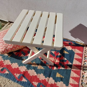 May include: White wooden folding table with a slatted top. The table is sitting on a colorful rug with fringe.