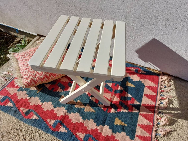 May include: White wooden folding table with a slatted top. The table is sitting on a colorful rug with fringe.
