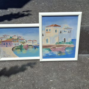 May include: Two framed paintings depicting coastal scenes with white buildings and small boats on blue water. The left painting shows a harbor with multiple boats, while the right painting features a single pink boat near white architecture.
