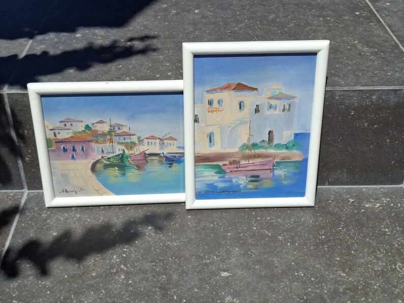May include: Two framed paintings depicting coastal scenes with white buildings and small boats on blue water. The left painting shows a harbor with multiple boats, while the right painting features a single pink boat near white architecture.