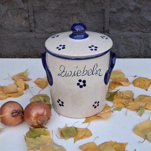 May include: A white ceramic onion storage jar with a blue lid and blue floral accents. The jar has a blue inscription that reads "Zwiebeln".