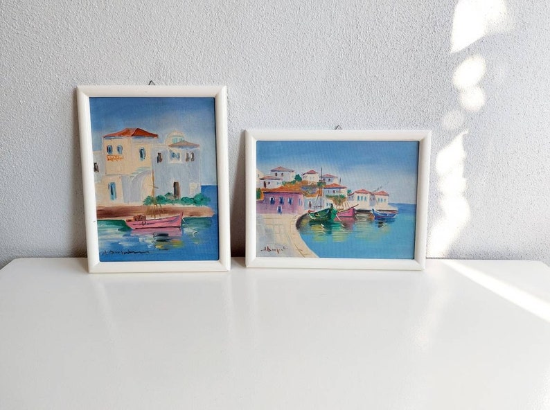 May include: Two framed oil paintings depicting coastal Mediterranean scenes with white buildings, red roofs, and colorful fishing boats floating on blue water. Each artwork is enclosed in a simple white frame with a small metal hanging loop at the top.