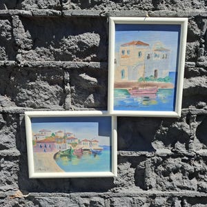 May include: Two framed paintings featuring coastal scenes with white buildings and small boats on blue water. The top painting shows a single pink boat near a building, while the bottom painting depicts a harbor with multiple colorful boats and houses.
