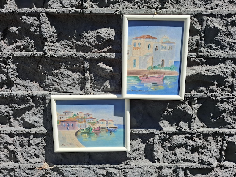 May include: Two framed paintings featuring coastal scenes with white buildings and small boats on blue water. The top painting shows a single pink boat near a building, while the bottom painting depicts a harbor with multiple colorful boats and houses.