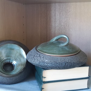 May include: Two ceramic pieces, a bowl and a lidded container, sit on a shelf. The bowl is gray with a teal interior. The lidded container is gray with a green lid and handle. The items rest on a stack of books and a light blue cloth.