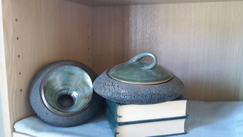 May include: Two ceramic pieces, a bowl and a lidded container, sit on a shelf. The bowl is gray with a teal interior. The lidded container is gray with a green lid and handle. The items rest on a stack of books and a light blue cloth.