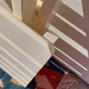 May include: White wooden shelf unit with a simple design. The shelf is made of multiple slats and has a white finish. The shelf is angled and the top edge is slightly chipped.