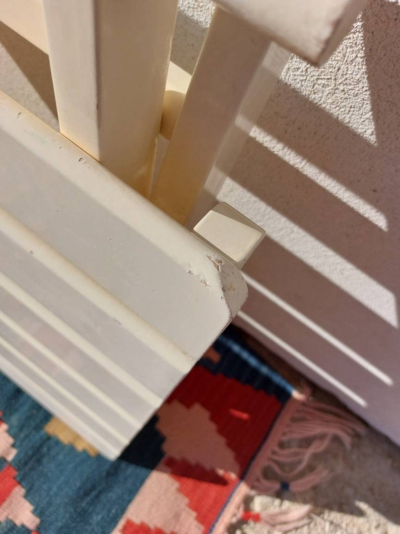 May include: White wooden shelf unit with a simple design. The shelf is made of multiple slats and has a white finish. The shelf is angled and the top edge is slightly chipped.