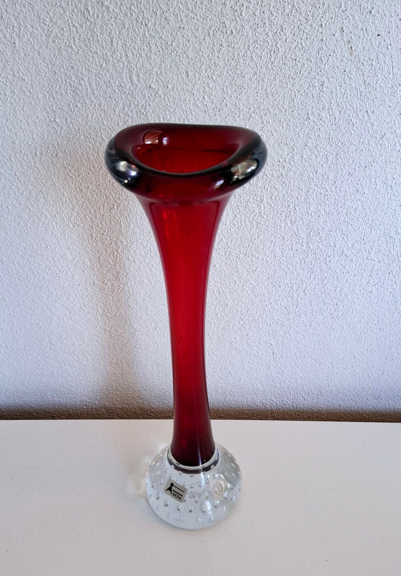 Peut inclure: Un vase en verre rouge et haut avec une ouverture &eacute;vas&eacute;e et un pied clair textur&eacute;. Le pied porte une petite &eacute;tiquette indiquant "Aalto 1962".
