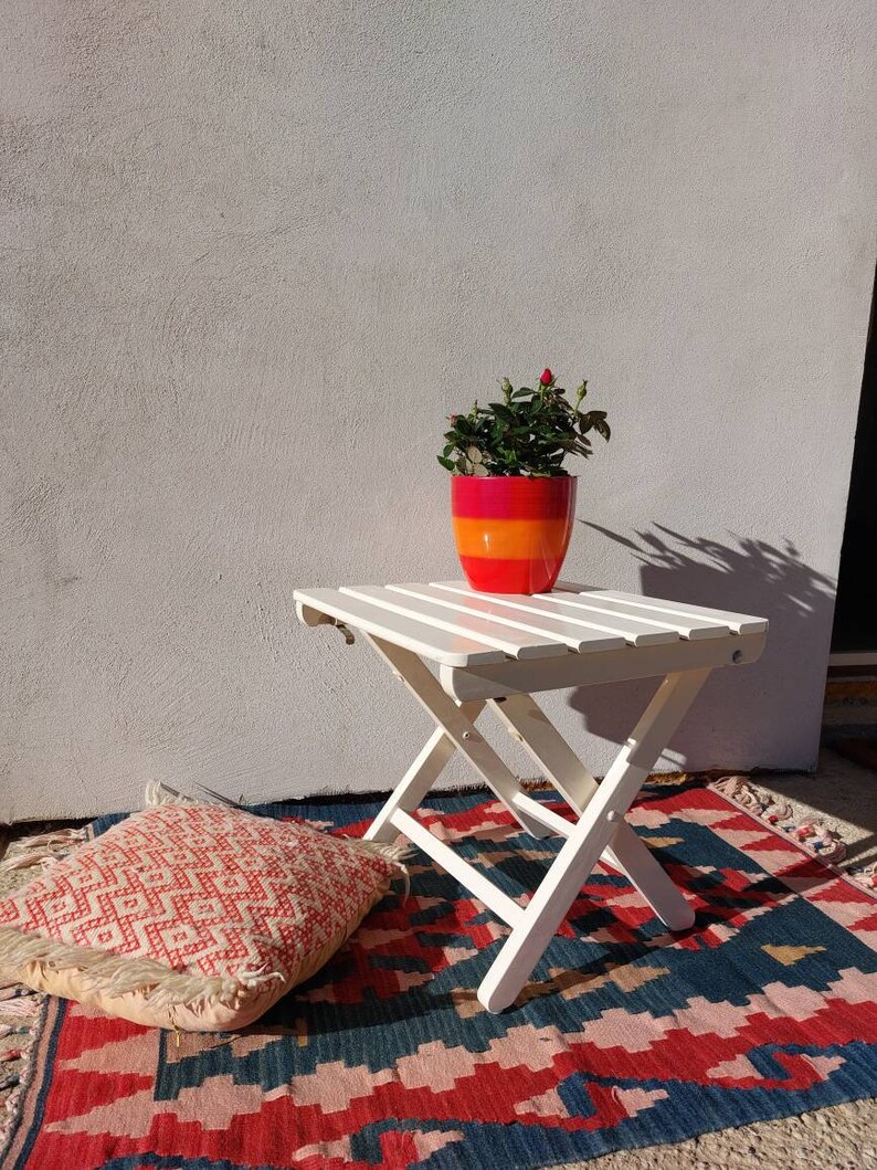 May include: White folding table with a red and orange planter with a small green plant on top. The table is on a rug with red, blue, and white patterns. A pink and white pillow is on the rug.