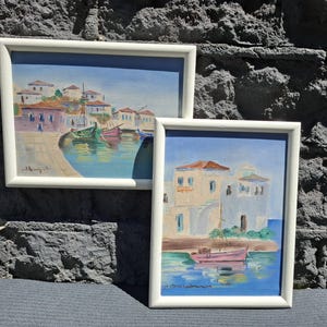 May include: Two framed paintings featuring coastal scenes with white buildings and colorful boats on blue water. The larger landscape painting shows a harbor with multiple boats, while the smaller portrait painting depicts a single pink boat.