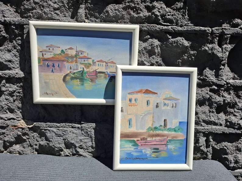May include: Two framed paintings featuring coastal scenes with white buildings and colorful boats on blue water. The larger landscape painting shows a harbor with multiple boats, while the smaller portrait painting depicts a single pink boat.